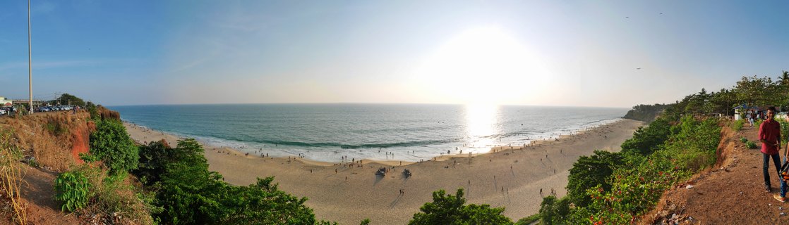 Panoramic coastal landscape showing Varkala Beach with red cliffs on both sides, sandy shore with people, and sun reflecting on the Arabian Sea