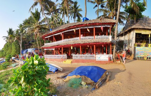 Two-story beachside restaurant with thatched roof, red columns, and white railings surrounded by palm trees on sandy beach in Varkala, India, with fishing equipment and blue tarps in foreground