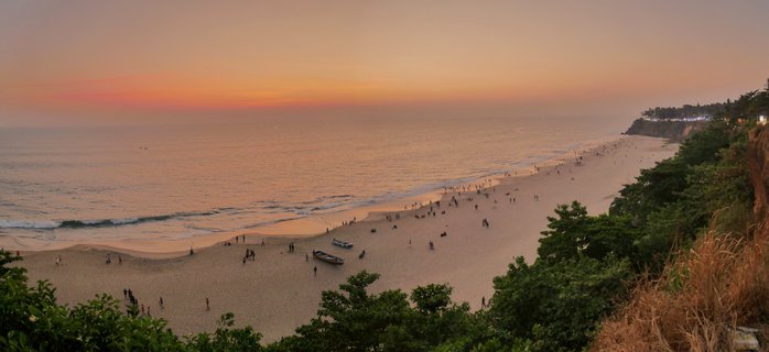 Aerial view of Varkala Beach at sunset showing orange-pink sky over the Arabian Sea, sandy beach with scattered people and fishing boats, green vegetation and cliff with lights on the right side