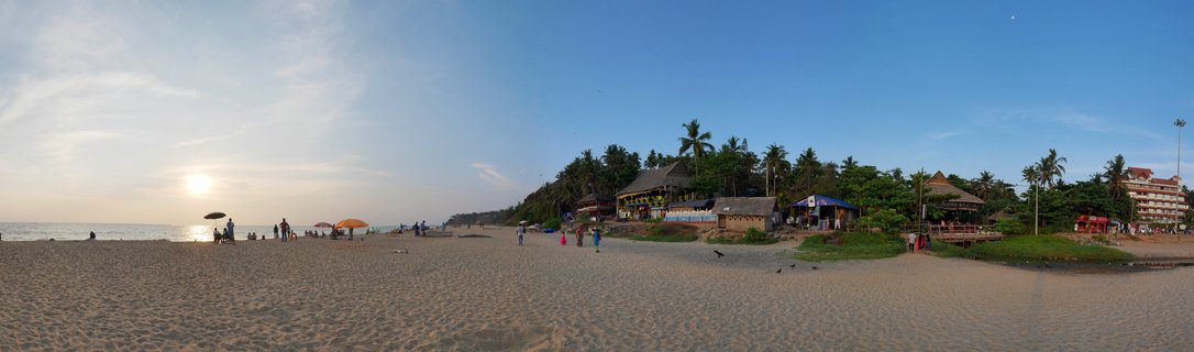 Wide beach scene at sunset showing golden sand, ocean on the left, people walking, orange beach umbrellas, palm trees, thatched-roof beach shacks, and a hotel building under blue sky with the setting sun visible