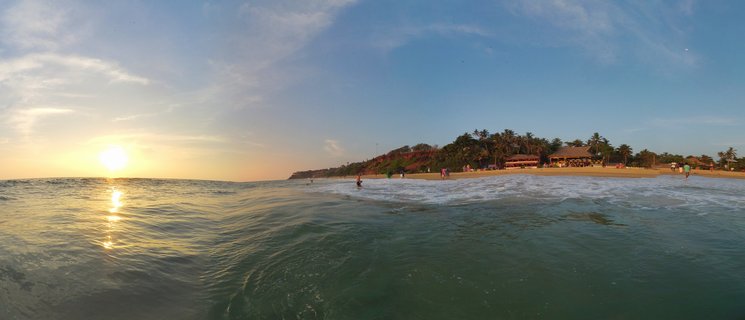Sunset over ocean at Varkala Beach with golden sun reflection on water waves, sandy shoreline with people, red cliffs and palm trees in background under blue sky with clouds