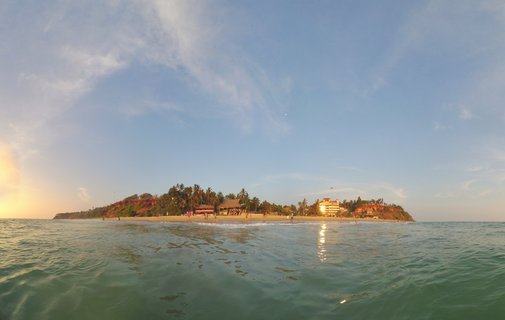 Wide beach scene showing Varkala's red cliffs and palm trees from water level during sunset with people on the beach and buildings along the shoreline under a blue sky with wispy clouds