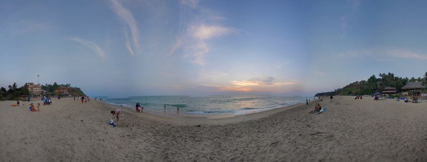Wide beach scene at Varkala, Kerala showing sand, ocean waves, scattered people, cliffs with buildings and palm trees on both sides, and a blue sky with wispy clouds at sunset