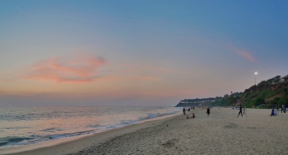 Wide beach scene at sunset showing ocean waves on left, sandy shore with scattered people walking and sitting, cliff with vegetation on right, pink clouds in blue sky