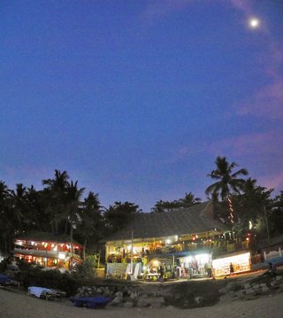 Beach shacks with warm lighting at twilight on Varkala Beach, Kerala, India, with palm trees silhouetted against a deep blue sky with visible moon