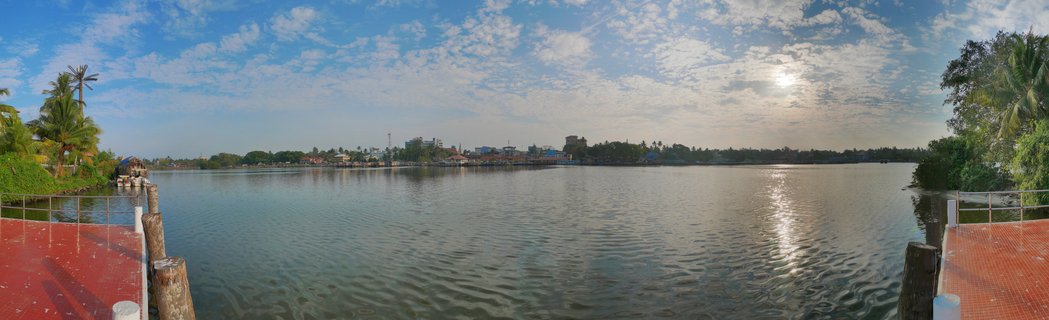 Wide panoramic view of Varkala backwaters in Kerala, India showing calm water reflecting sunlight, buildings along the distant shore, palm trees on both sides, and red-tiled platforms in the foreground under a blue sky with scattered clouds