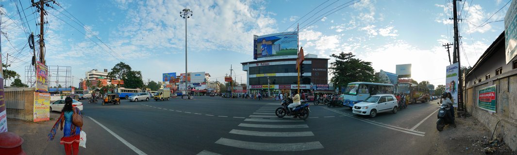 Wide street intersection in Varkala, India showing vehicles including auto-rickshaws, motorcycles, buses and cars, with pedestrians, commercial buildings with billboards, power lines, and blue sky with scattered clouds