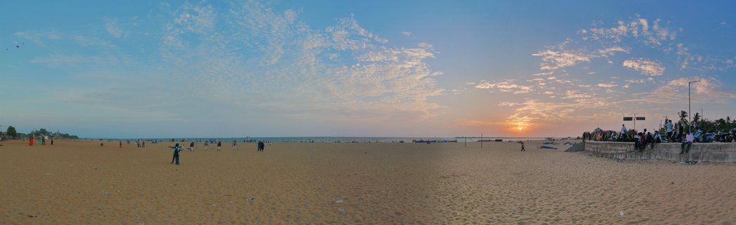 Wide beach scene at sunset showing golden sand, ocean horizon, scattered people walking and sitting on a seawall, with orange and blue sky filled with clouds