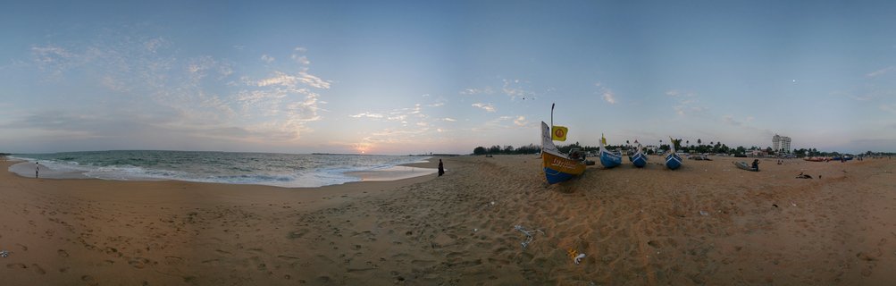Panoramic view of Varkala Beach in Kerala, India showing traditional blue and yellow fishing boats on sand, Arabian Sea waves, palm trees, distant white hotel building, and sunset sky with scattered clouds