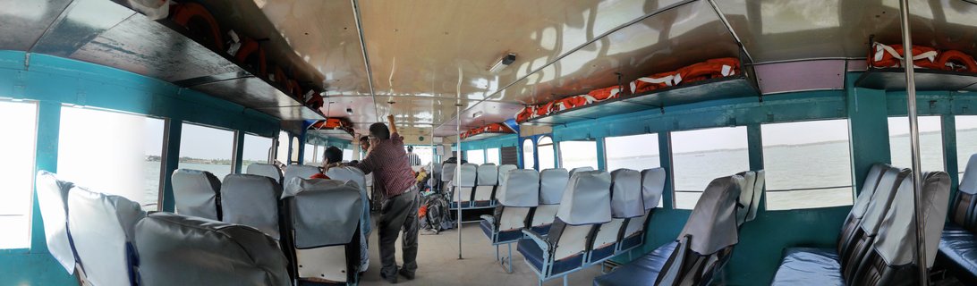 Interior view of a passenger ferry boat showing rows of blue and grey vinyl seats, teal-painted walls and ceiling, orange life jackets stored on overhead shelves, metal support poles, and passengers visible near the front with water visible through windows on both sides