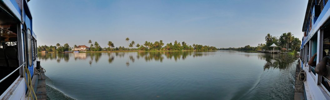 Panoramic view from a boat showing calm backwaters in Kerala, India with coconut palm trees lining the shore and traditional buildings with red-tiled roofs visible in the distance under a clear blue sky