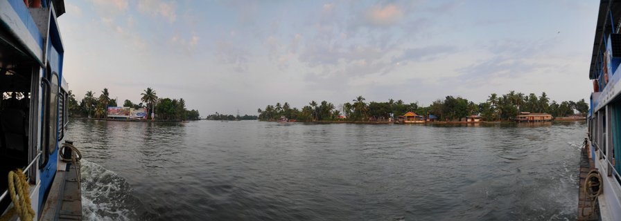 Wide panoramic photograph of Kerala backwaters showing calm water with houseboats and buildings along tree-lined shores, viewed from a blue boat with visible wake in the foreground