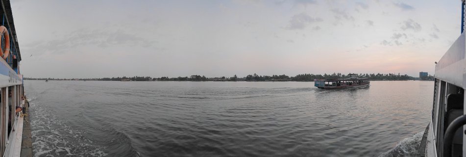 View from a boat showing a passenger ferry on Ashtamudi Lake with palm tree-lined shore and cloudy sky at dusk in Kollam, Kerala, India