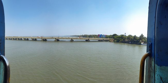 Long concrete bridge spanning across calm greenish-brown water with palm trees on the far shore, viewed through blue metal boat frame