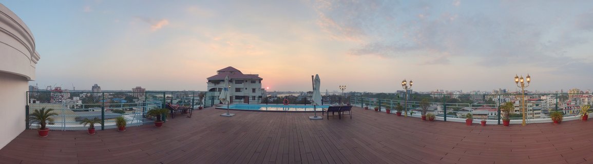 Panoramic view of a rooftop swimming pool deck with wooden flooring, potted plants, and lounge chairs overlooking Kochi city buildings at sunset