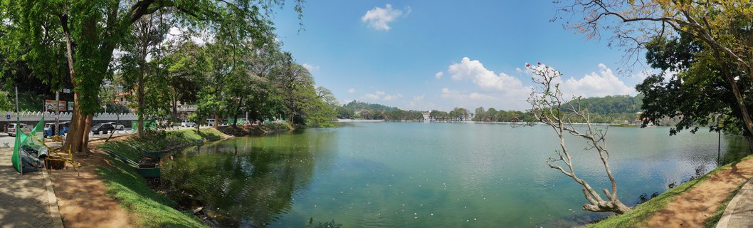 Wide panoramic photograph of Kandy Lake in Sri Lanka showing calm green water, tree-lined shores, paved walkway with people, parked vehicles, distant hills, and blue sky with white clouds
