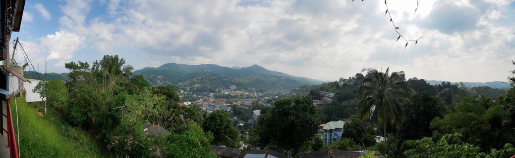 Panoramic landscape showing Kandy city buildings spread across green mountain hills in Sri Lanka, with tropical trees, palm trees, cloudy sky, and distant mountain ranges visible in the background