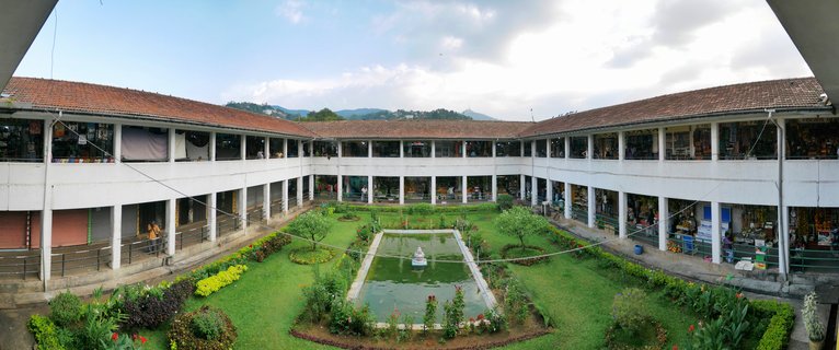 White two-story building complex with red tiled roofs surrounding a green courtyard with rectangular pond and fountain in Kandy Sri Lanka
