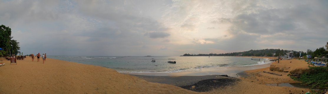 Wide beach scene showing golden sand curving around a calm bay with several small boats anchored in turquoise water, people walking along the shore, palm trees on the left, buildings on the right, and cloudy sky with sun breaking through near the horizon