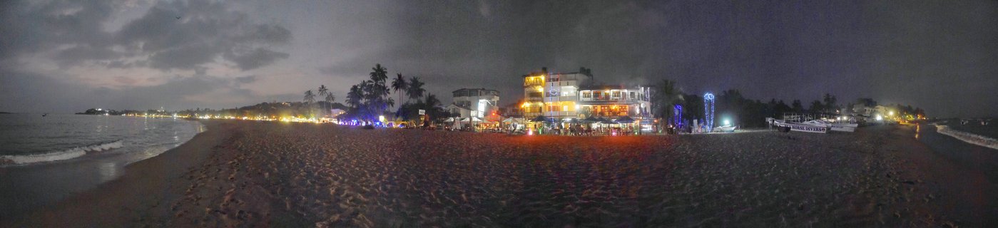 Panoramic view of Unawatuna Beach at twilight showing illuminated restaurants and buildings along the shoreline, palm trees, boats on sand, and ocean waves under a cloudy evening sky