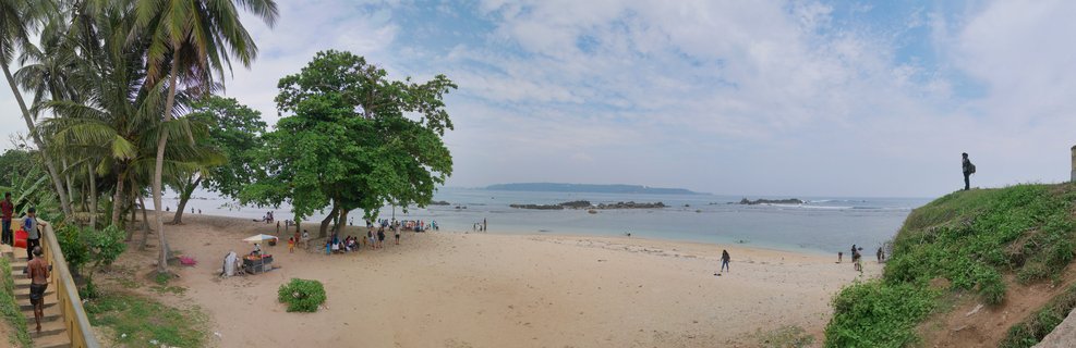 Panoramic beach view showing palm trees, people on sand, calm ocean water with rocks, and cloudy sky in Galle, Sri Lanka