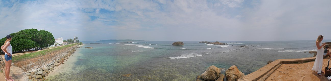 Panoramic view of Galle Fort stone walls along the coast with turquoise ocean water, rocky outcrops, colonial buildings, and visitors on the ramparts in Sri Lanka