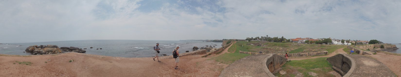 Two people walking on a dirt path along historic stone fortification walls with ocean view, rocky coastline, grass areas, and colonial buildings with red roofs in the distance under cloudy sky
