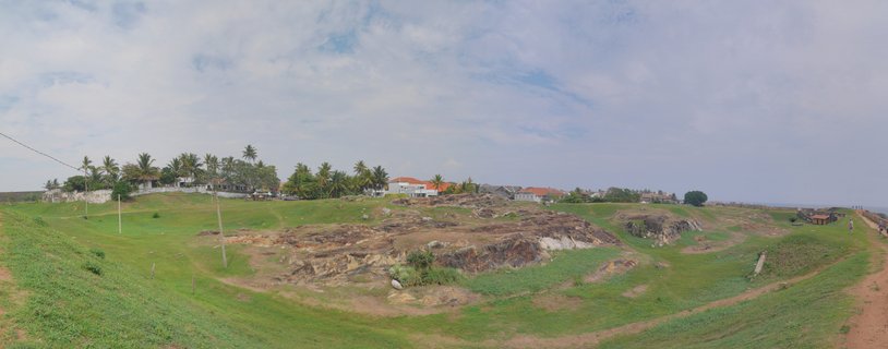 Panoramic coastal landscape showing green grassy terrain with rocky outcrops, palm trees, white buildings with red roofs, and ocean visible in the distance under cloudy sky