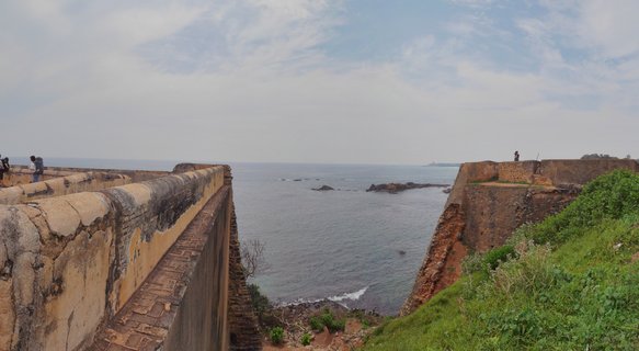 Stone fortification walls of Galle Fort extending toward the ocean with people walking on the ramparts, green vegetation on the right side, and cloudy sky above the Indian Ocean coastline