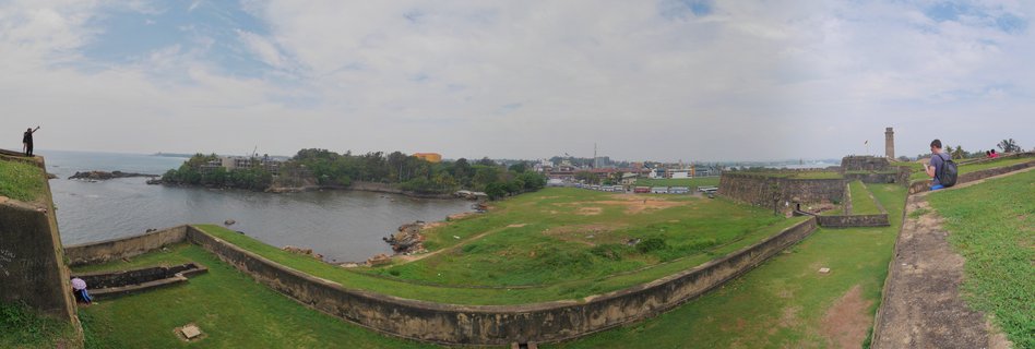 Panoramic photograph showing the historic stone walls and grassy grounds of Galle Fort in Sri Lanka, with the ocean visible on the left and the lighthouse tower on the right under a cloudy sky