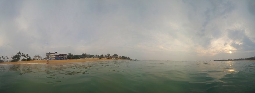 Split-level photograph showing ocean water in foreground and beach with buildings and palm trees along the horizon at sunset in Galle, Sri Lanka