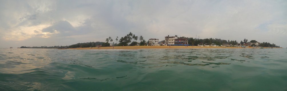 Panoramic water-level view of Galle Beach showing traditional wooden fishing boats on golden sand with coconut palm trees and white-blue coastal buildings along the shoreline, calm turquoise ocean water in foreground, overcast sky with light breaking through clouds