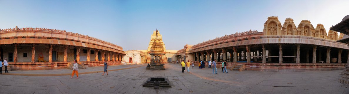 Wide panoramic view of ancient Hindu temple courtyard in Hampi India showing stone pillared corridors on both sides with central shrine tower and visitors walking on paved ground under clear blue sky