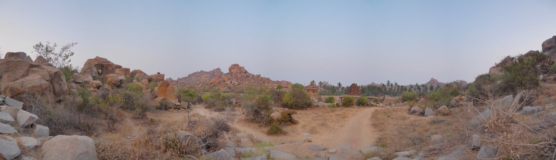 Panoramic landscape showing ancient temple ruins among large granite boulders and rocky hills in Hampi, India, with dirt paths and sparse vegetation under a clear sky