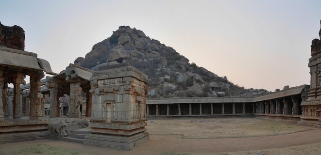Ancient stone temple ruins with carved pillars surrounding an open courtyard, with a large rocky hill covered in boulders rising in the background under a pale sky