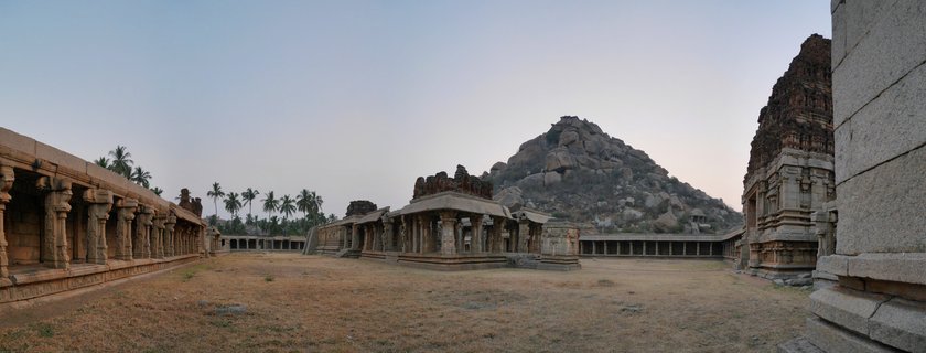 Ancient stone temple ruins with carved pillars and pavilions in a courtyard setting, with a rocky hill and palm trees in the background at Hampi, India
