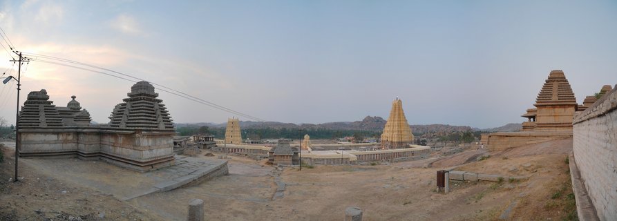 Panoramic view of ancient stone Hindu temples with pyramidal towers at Hampi archaeological site, with rocky hills in the background under a pale evening sky