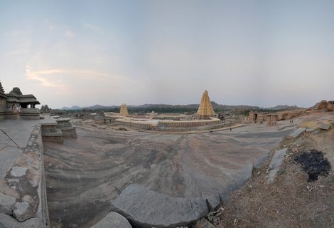 Wide view of Virupaksha Temple complex showing the main gopuram tower and ancient stone structures set against rocky granite terrain and distant hills under a pale evening sky at Hampi UNESCO site