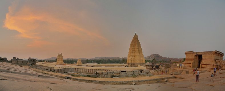 Ancient stone temple ruins with pyramidal tower structures on rocky ground at sunset in Hampi, India, with warm orange clouds in the sky and visitors walking near the structures