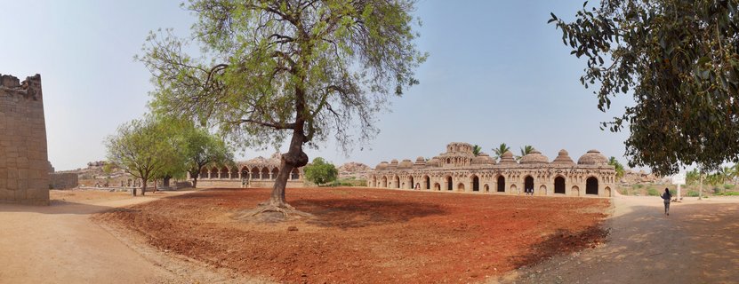 Historic stone building with multiple domed chambers and arched doorways at Hampi archaeological site, with large tree in foreground on reddish earth under clear sky
