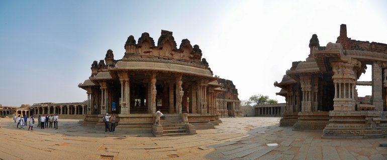 Ancient stone temple structures with carved pillars and ornate roof details in a courtyard complex, with visitors walking on stone pavement under clear blue sky in Hampi, India