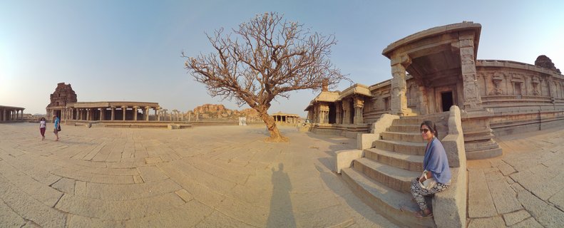 Wide view of ancient stone temple ruins with carved pillars, paved courtyard, bare tree in center, and visitors exploring the historic Hampi UNESCO site in India