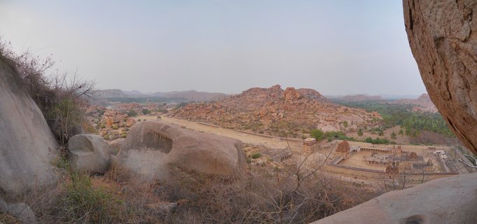 Panoramic landscape showing ancient temple ruins surrounded by large granite boulders and rocky hills in Hampi, India, with sparse vegetation and palm trees visible in the valley below
