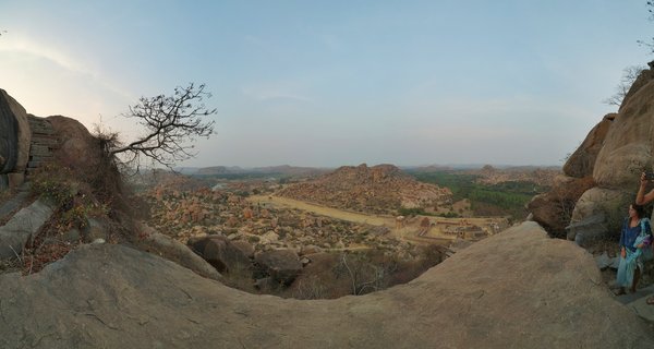 Panoramic landscape showing ancient temple ruins among scattered boulders in Hampi, India, with a lone tree on rocky foreground and hills in the distance under clear sky