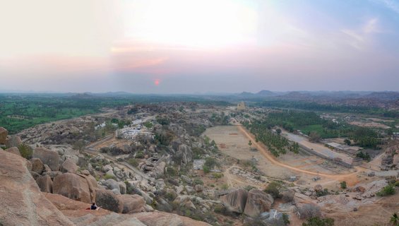 Panoramic sunset view over Hampi showing ancient temple towers, large boulder formations, green agricultural fields, and palm trees in Karnataka, India