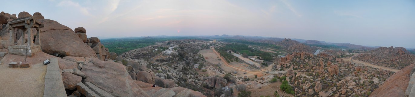 Wide panoramic landscape showing ancient stone temple structure on rocky outcrop overlooking Hampi valley with boulder formations, sparse vegetation, and setting sun on horizon