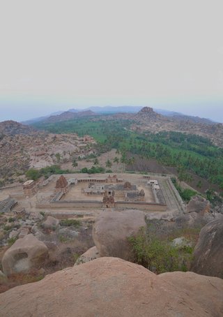 Aerial photograph of ancient stone temple ruins with tower structures enclosed by walls, surrounded by large granite boulders and palm trees in Hampi, India