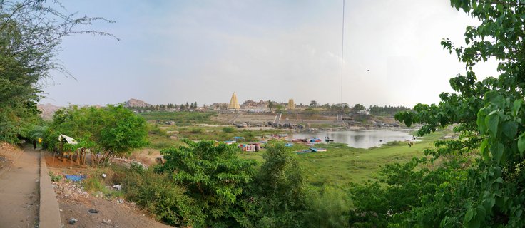 Wide landscape showing Hindu temple towers along a river with green trees in foreground and boats on the water in Hampi, India