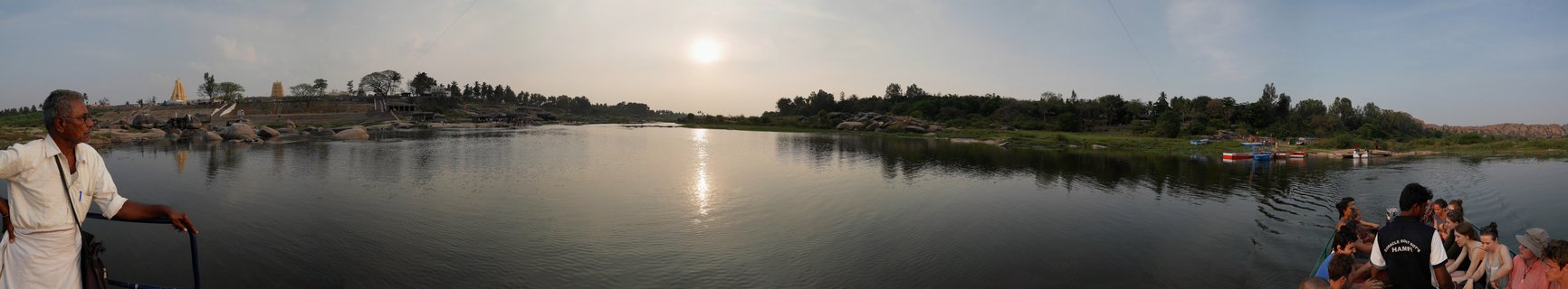 Wide river view showing a man in white clothing on the left, tourists in a round coracle boat on the right, and ancient temple structures on the rocky far bank under a golden sky with sun reflection on water