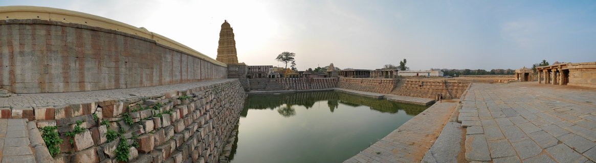 Ancient stone temple tank with stepped ghats surrounding still green water, with a tall temple tower visible in the background at Hampi, India